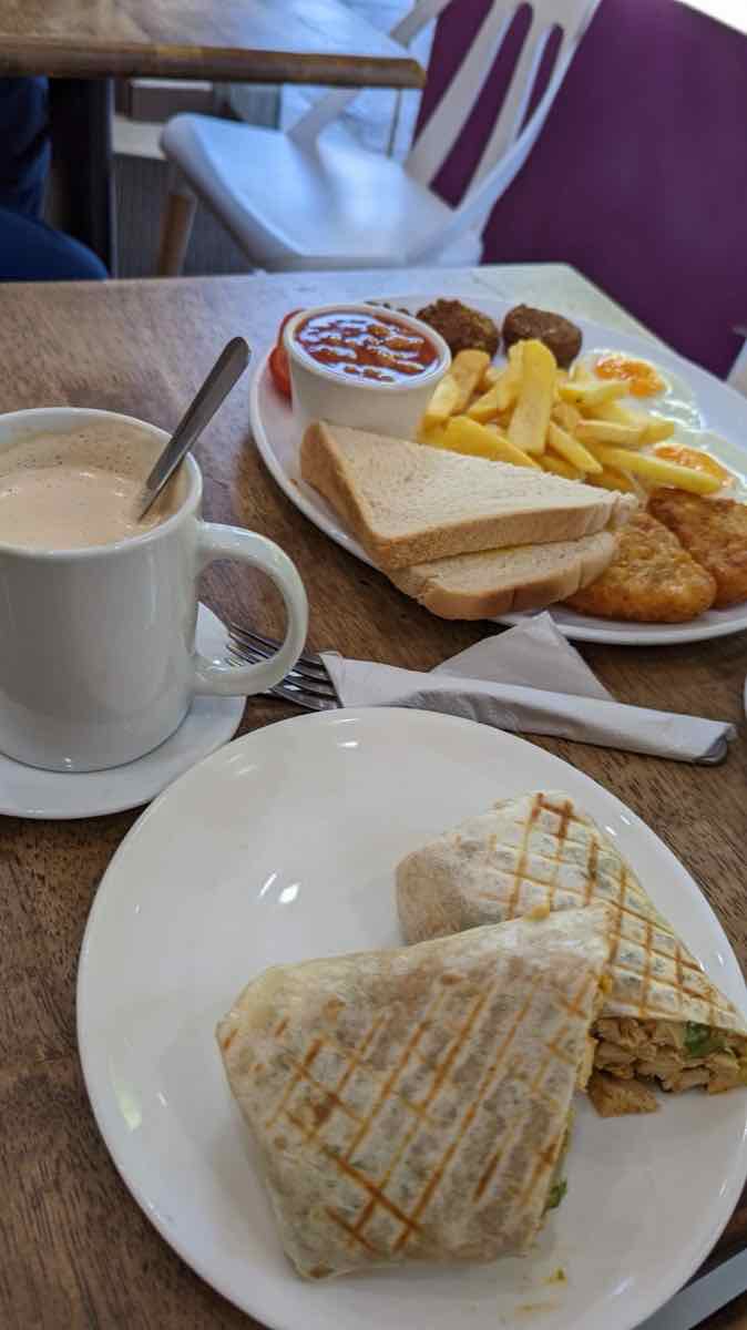 Griddled chicken wrap on a plate with a mug of tea and a breakfast behind